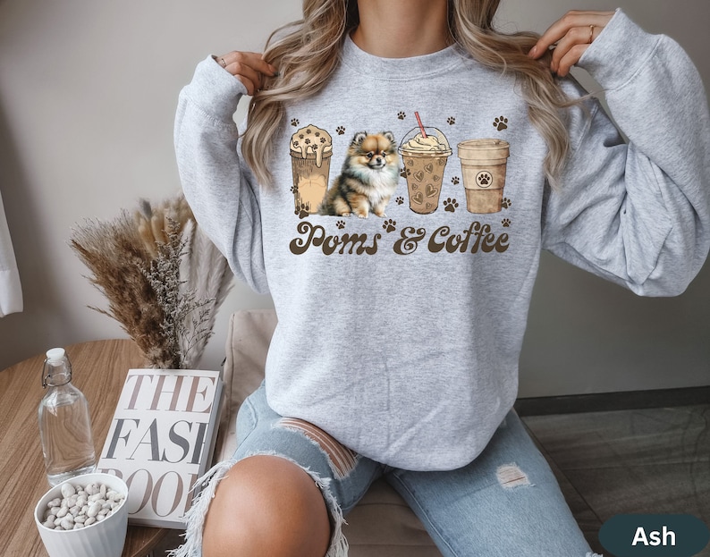 Puede incluir: Sudadera gris con un gr&aacute;fico de un perro Pomerania y tres tazas de caf&eacute;. El texto "Poms & Coffee" est&aacute; impreso en la sudadera.