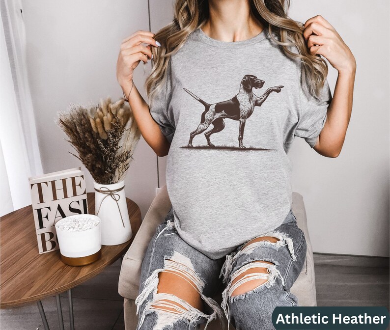 Pu&ograve; includere: Una maglietta grigia con un'illustrazione in bianco e nero di un Bracco tedesco a pelo corto che punta. La maglietta &egrave; etichettata "Athletic Heather".