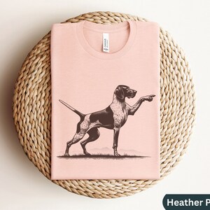 Pu&ograve; includere: Una maglietta rosa con un'illustrazione in bianco e nero di un Bracco tedesco a pelo corto che punta. Il testo "Heather Peach" &egrave; visibile nell'angolo in basso a destra dell'immagine.