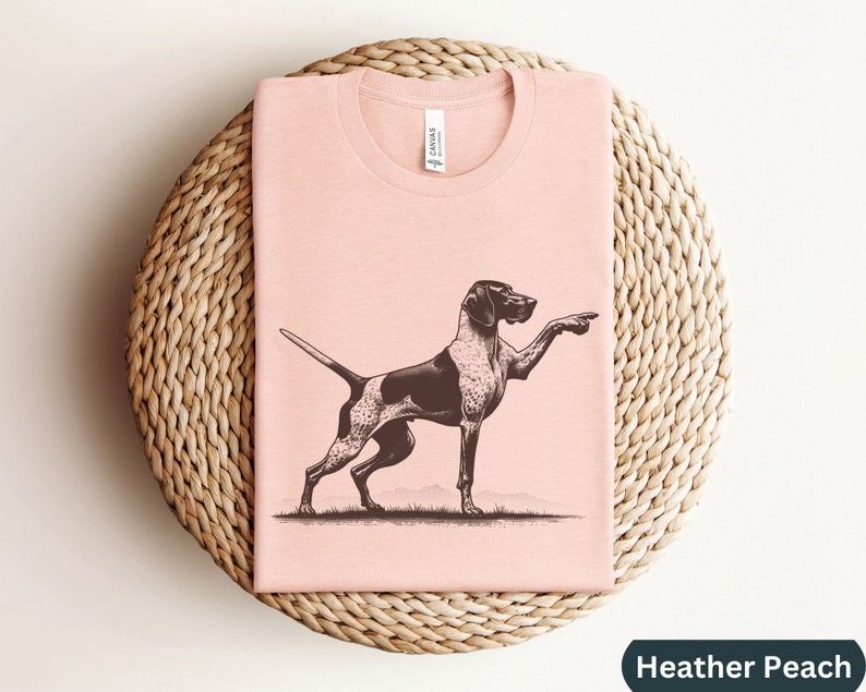 Pu&ograve; includere: Una maglietta rosa con un'illustrazione in bianco e nero di un Bracco tedesco a pelo corto che punta. Il testo "Heather Peach" &egrave; visibile nell'angolo in basso a destra dell'immagine.