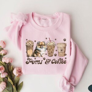 Puede incluir: Sudadera rosa con un gr&aacute;fico marr&oacute;n de un perro pomerania, dos tazas de caf&eacute; y el texto "Poms & Coffee".