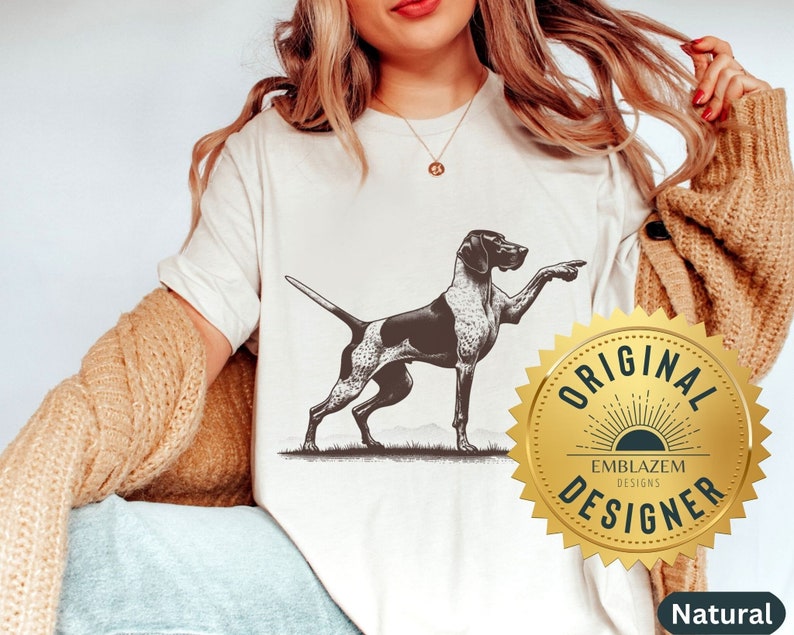 Pu&ograve; includere: Una maglietta bianca con un'illustrazione in bianco e nero di un cane che punta a destra. La maglietta ha un sigillo dorato con il testo "Original Emblazem Designs Designer" su di esso.