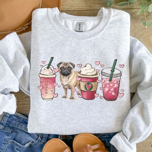 May include: A gray sweatshirt with a pug dog illustration and three coffee cups with hearts. The cups are filled with pink and white beverages. The text "Ash" is visible in the bottom right corner.