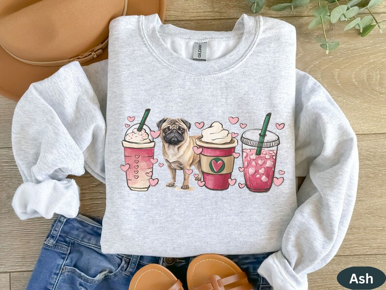 May include: A gray sweatshirt with a pug dog illustration and three coffee cups with hearts. The cups are filled with pink and white beverages. The text "Ash" is visible in the bottom right corner.