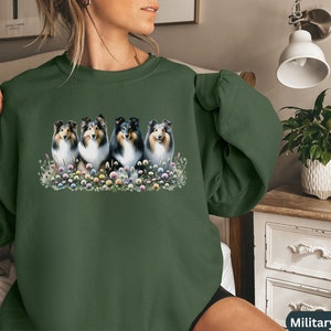 Puede incluir: Una sudadera verde con un gr&aacute;fico de cuatro perros Collie en un campo de flores. Los perros son negros y blancos con ojos marrones. Las flores son rosas, moradas y amarillas.