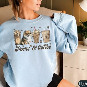 Puede incluir: Sudadera azul claro con un gr&aacute;fico de un perro Pomerania y cuatro tazas de caf&eacute;. El texto "Poms & Coffee" est&aacute; impreso en la sudadera.