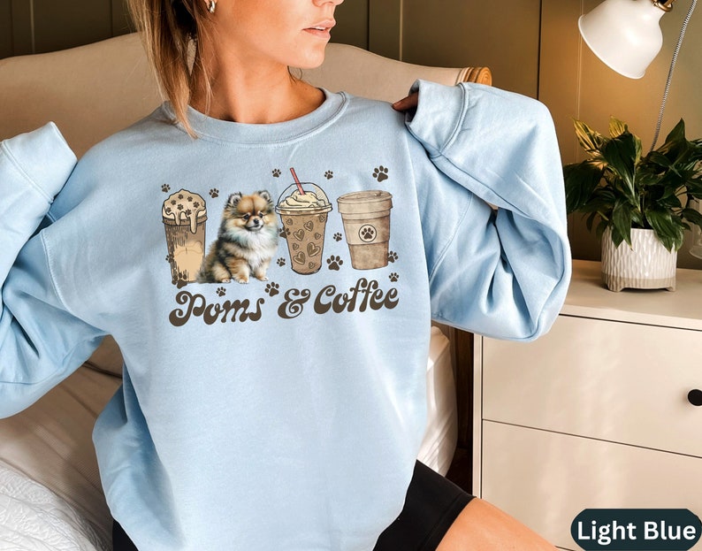 Puede incluir: Sudadera azul claro con un gr&aacute;fico de un perro Pomerania y cuatro tazas de caf&eacute;. El texto "Poms & Coffee" est&aacute; impreso en la sudadera.