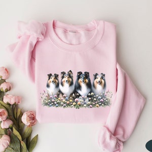 Puede incluir: Sudadera rosa con un gr&aacute;fico de cuatro Shetland Sheepdogs sentados en un campo de flores.