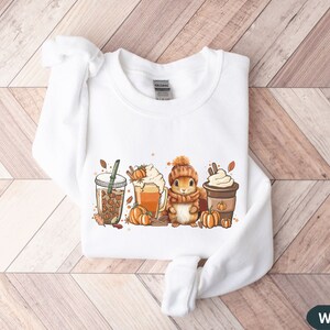 May include: White sweatshirt with a graphic of a squirrel holding a cup of coffee, surrounded by fall-themed elements like pumpkins, leaves, and coffee cups.