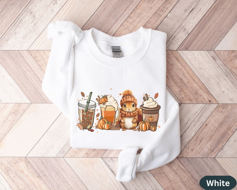 May include: White sweatshirt with a graphic of a squirrel holding a cup of coffee, surrounded by fall-themed elements like pumpkins, leaves, and coffee cups.