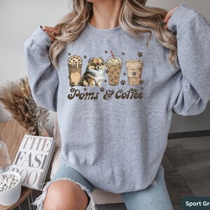 Puede incluir: Una sudadera gris con un gr&aacute;fico de un perro Pomerania y tres tazas de caf&eacute;. El texto "Poms & Coffee" est&aacute; impreso en la sudadera. La sudadera est&aacute; disponible en Gris Deportivo.