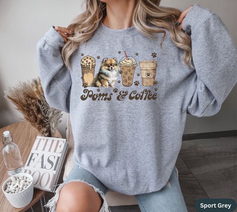 Puede incluir: Una sudadera gris con un gr&aacute;fico de un perro Pomerania y tres tazas de caf&eacute;. El texto "Poms & Coffee" est&aacute; impreso en la sudadera. La sudadera est&aacute; disponible en Gris Deportivo.