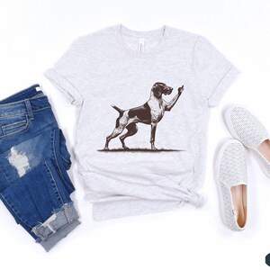 May include: A gray t-shirt with a black and white illustration of a dog giving the middle finger. The shirt is on a white background with a pair of white shoes and a pair of blue jeans.