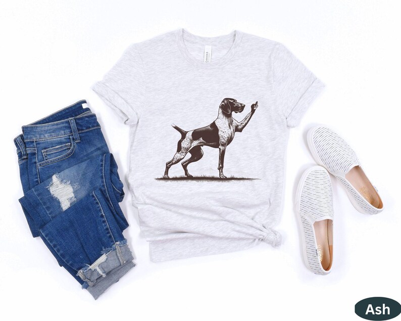 May include: A gray t-shirt with a black and white illustration of a dog giving the middle finger. The shirt is on a white background with a pair of white shoes and a pair of blue jeans.