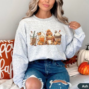May include: A gray sweatshirt with a graphic of a squirrel holding a cup of coffee, surrounded by fall-themed imagery like pumpkins, leaves, and coffee cups. The sweatshirt has a crew neck and long sleeves.