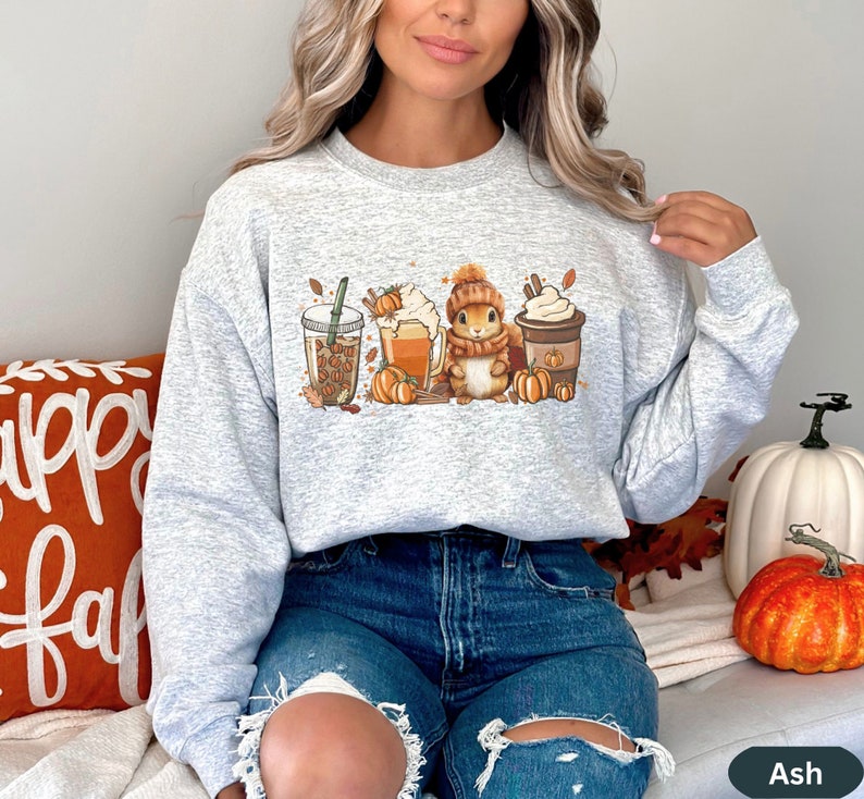 May include: A gray sweatshirt with a graphic of a squirrel holding a cup of coffee, surrounded by fall-themed imagery like pumpkins, leaves, and coffee cups. The sweatshirt has a crew neck and long sleeves.
