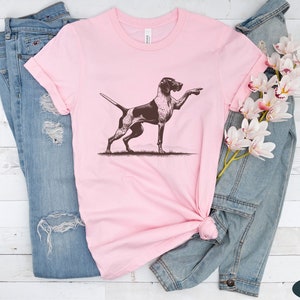 Pu&ograve; includere: Una maglietta rosa con un'illustrazione marrone e bianca di un Bracco tedesco a pelo corto che punta con la zampa destra.