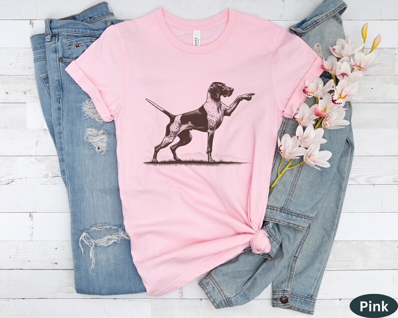 Pu&ograve; includere: Una maglietta rosa con un'illustrazione marrone e bianca di un Bracco tedesco a pelo corto che punta con la zampa destra.