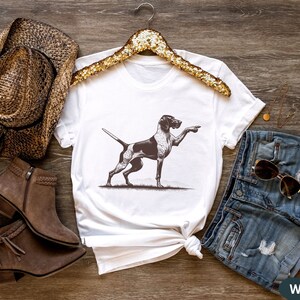 Pu&ograve; includere: Maglietta bianca con un'illustrazione in bianco e nero di un cane che punta. Il cane &egrave; su uno sfondo bianco. La maglietta &egrave; su una superficie di legno con un cappello da cowboy marrone, stivali marroni e pantaloncini di jeans blu.