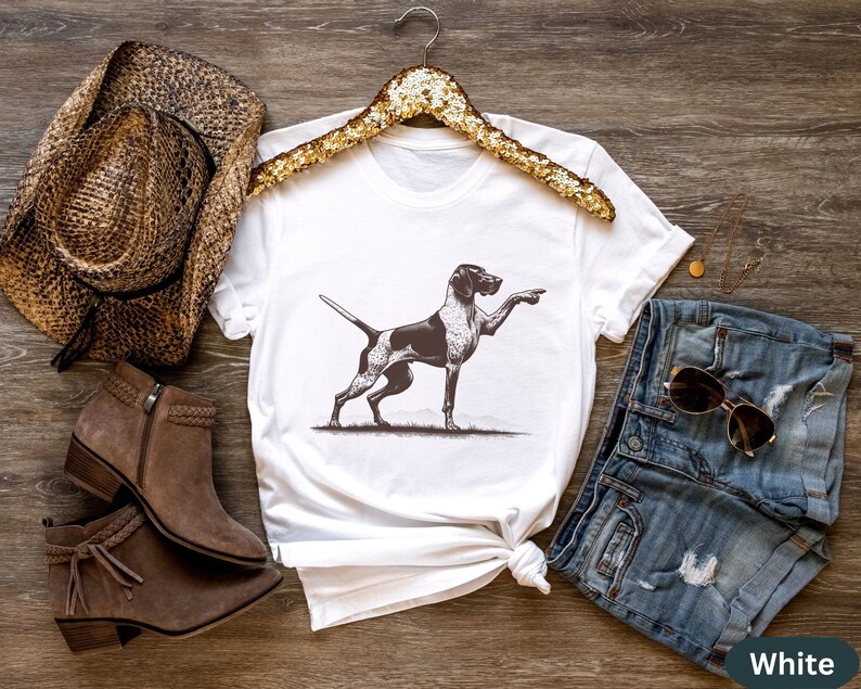 Pu&ograve; includere: Maglietta bianca con un'illustrazione in bianco e nero di un cane che punta. Il cane &egrave; su uno sfondo bianco. La maglietta &egrave; su una superficie di legno con un cappello da cowboy marrone, stivali marroni e pantaloncini di jeans blu.