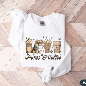 Puede incluir: Sudadera blanca con un perro pomerania marr&oacute;n, dos vasos de caf&eacute; helado y el texto "Poms & Coffee".