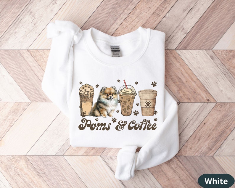 Puede incluir: Sudadera blanca con un perro pomerania marr&oacute;n, dos vasos de caf&eacute; helado y el texto "Poms & Coffee".