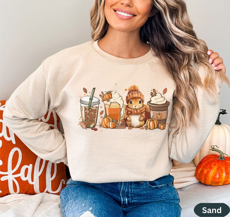 May include: A beige sweatshirt with a graphic print of a squirrel, a cup of coffee, a glass of iced coffee, and a pumpkin spice latte. The graphic also includes fall leaves and pumpkins.