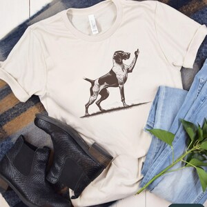 May include: A white t-shirt with a black and white illustration of a German Shorthaired Pointer dog giving a thumbs up. The dog is standing on a brown patch of grass. The shirt is laying on a brown and white striped blanket with a pair of black leather boots and blue jeans.