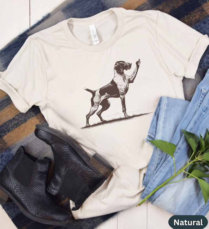 May include: A white t-shirt with a black and white illustration of a German Shorthaired Pointer dog giving a thumbs up. The dog is standing on a brown patch of grass. The shirt is laying on a brown and white striped blanket with a pair of black leather boots and blue jeans.