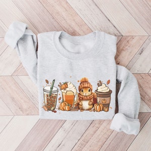 May include: A gray sweatshirt with a cute cartoon squirrel holding a cup of coffee and surrounded by fall-themed imagery, including pumpkins, leaves, and coffee cups.
