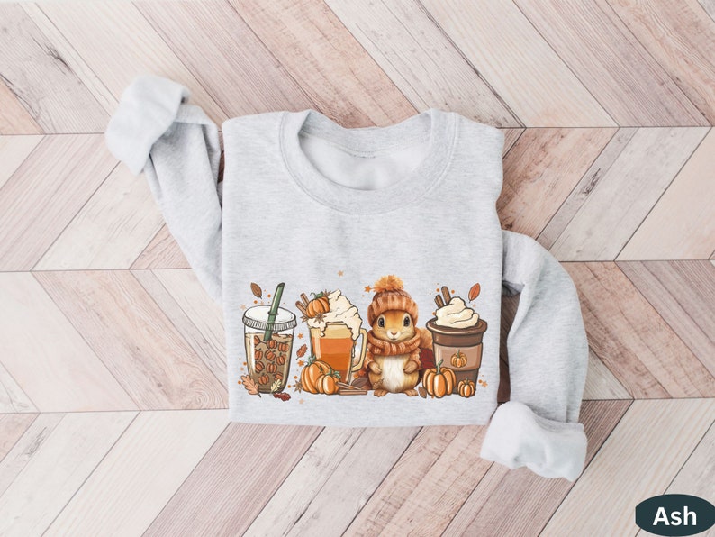 May include: A gray sweatshirt with a cute cartoon squirrel holding a cup of coffee and surrounded by fall-themed imagery, including pumpkins, leaves, and coffee cups.