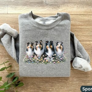Puede incluir: Una sudadera gris con un estampado de cinco perros Collie en un campo de flores. Los perros miran todos a la c&aacute;mara y tienen pelo negro y blanco.