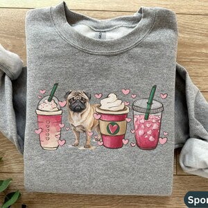 May include: A gray sweatshirt with a design of a pug dog and three coffee drinks with pink hearts. The drinks are a pink iced coffee, a caramel latte, and a regular iced coffee. The sweatshirt is labeled "Sport Grey".