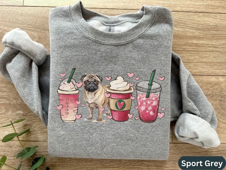 May include: A gray sweatshirt with a design of a pug dog and three coffee drinks with pink hearts. The drinks are a pink iced coffee, a caramel latte, and a regular iced coffee. The sweatshirt is labeled "Sport Grey".