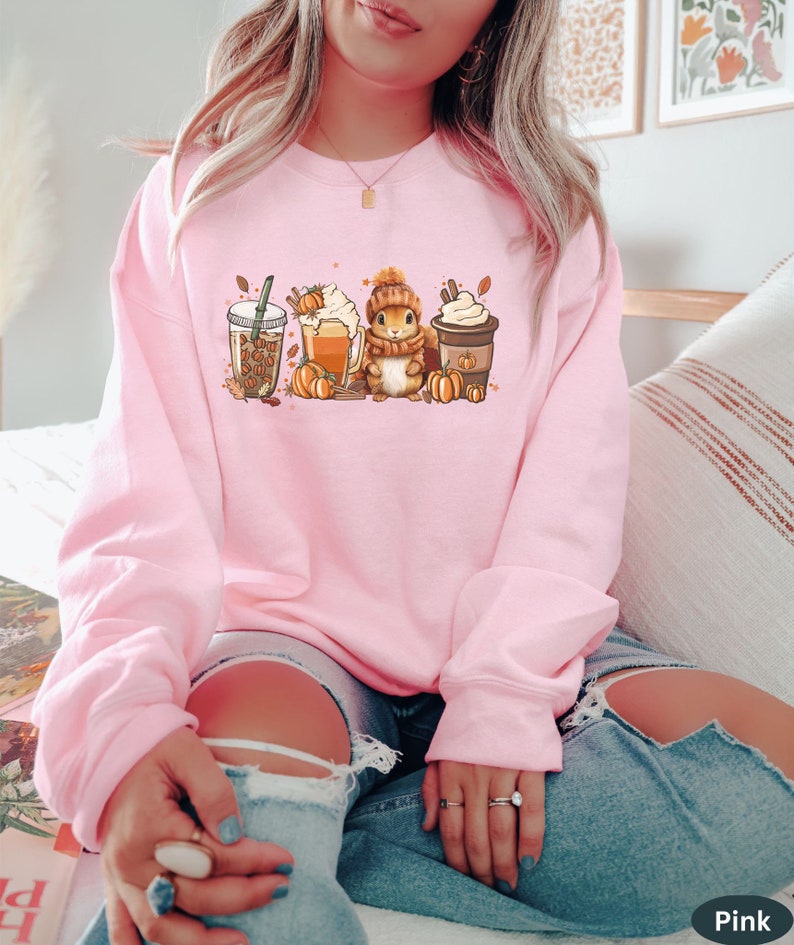 May include: A pink sweatshirt with a graphic of a squirrel holding a pumpkin spice latte, a pumpkin spice beer, and a pumpkin spice coffee. The graphic also includes fall leaves and pumpkins.
