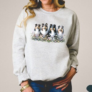 Puede incluir: Una sudadera gris con un gr&aacute;fico de cinco perros pastores Shetland sentados en un campo de flores. Los perros miran todos a la c&aacute;mara y tienen pelaje blanco y negro.