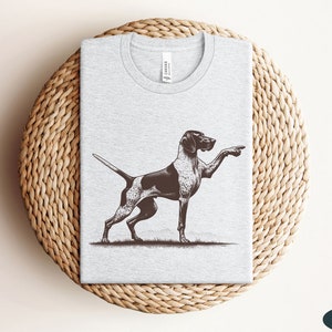 Pu&ograve; includere: Una maglietta grigia con un'illustrazione in bianco e nero di un Bracco tedesco a pelo corto che punta. Il cane &egrave; in piedi su un prato erboso. La maglietta ha un'etichetta che dice "Canvas".