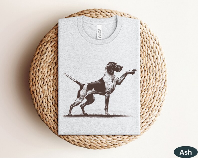 Pu&ograve; includere: Una maglietta grigia con un'illustrazione in bianco e nero di un Bracco tedesco a pelo corto che punta. Il cane &egrave; in piedi su un prato erboso. La maglietta ha un'etichetta che dice "Canvas".