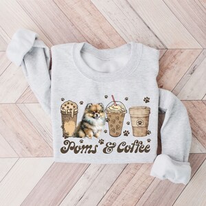 Puede incluir: Una sudadera gris con un lindo perro Pomerania y tres vasos de caf&eacute; helado. El texto "Poms & Coffee" est&aacute; impreso en la sudadera.
