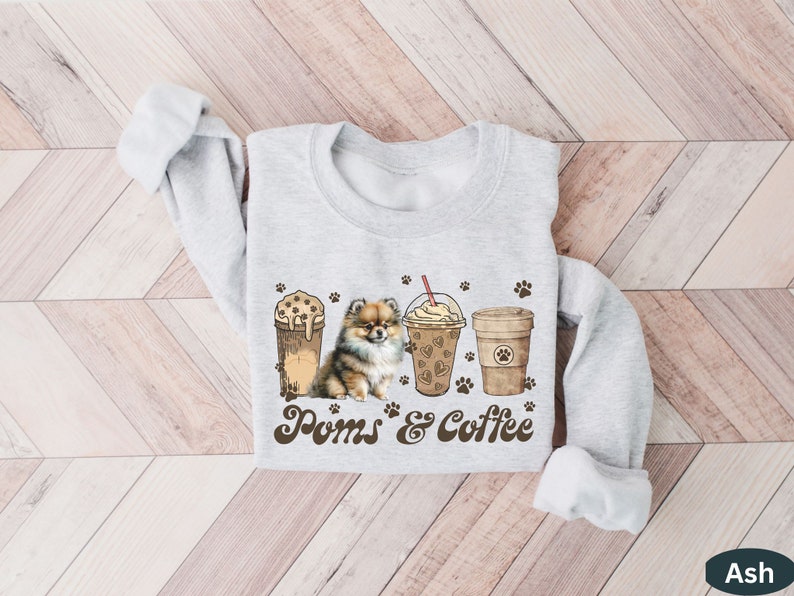 Puede incluir: Una sudadera gris con un lindo perro Pomerania y tres vasos de caf&eacute; helado. El texto "Poms & Coffee" est&aacute; impreso en la sudadera.