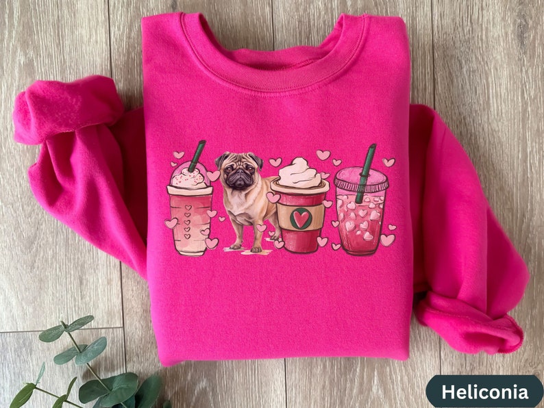 May include: Pink sweatshirt with a graphic of a pug dog standing in front of three coffee cups with hearts. The cups are filled with coffee, whipped cream, and iced coffee.