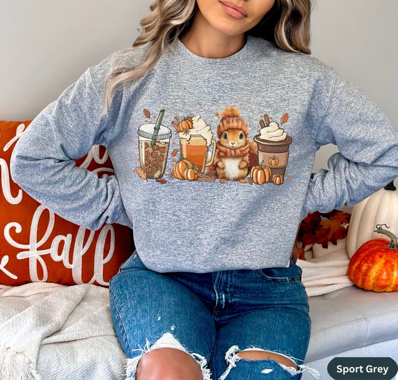 May include: A gray sweatshirt with a graphic of a squirrel holding a cup of coffee, surrounded by fall-themed elements like pumpkins, leaves, and coffee cups. The text "Sport Grey" is visible in the bottom right corner.