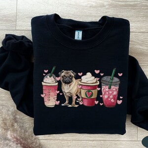 May include: Black sweatshirt with a pug dog and three pink and white coffee cups with hearts. The cups have green straws and are surrounded by pink hearts.