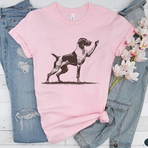 May include: A pink t-shirt with a black and white illustration of a German Shorthaired Pointer dog giving the middle finger.
