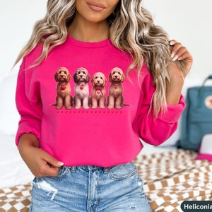 May include: A pink sweatshirt with a graphic of four brown and white doodle dogs wearing red hearts around their necks. The dogs are sitting in a row and there are small red hearts below them.