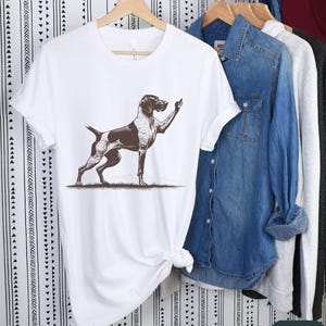 May include: White t-shirt with a brown and white German Shorthaired Pointer dog illustration. The dog is standing on its hind legs with its front paw raised in a gesture of giving the middle finger.