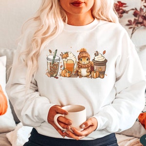May include: White sweatshirt with a graphic of a squirrel holding a cup of coffee, surrounded by fall-themed imagery like pumpkins, leaves, and coffee cups. The text "Fall Y'all" is printed on one of the coffee cups.