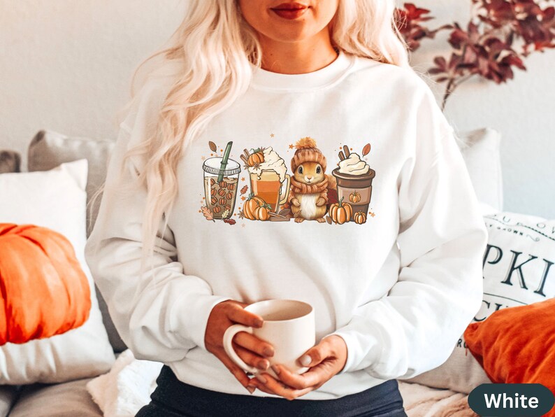 May include: White sweatshirt with a graphic of a squirrel holding a cup of coffee, surrounded by fall-themed imagery like pumpkins, leaves, and coffee cups. The text "Fall Y'all" is printed on one of the coffee cups.