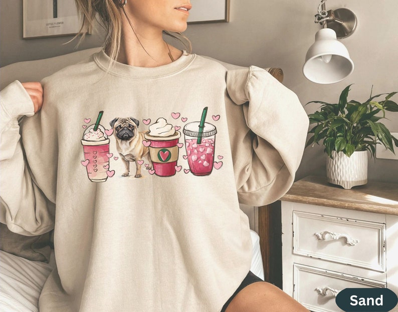 May include: A beige sweatshirt with a graphic design of a pug dog and four cups of coffee with hearts. The cups are decorated with pink and green accents.