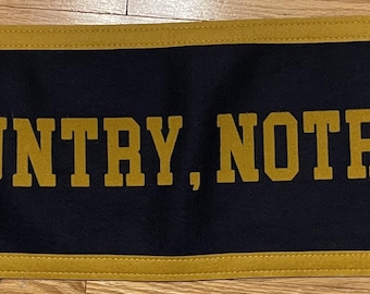 Może przedstawiać: Granatowy baner ze złotym napisem "GOD, COUNTRY, NOTRE DAME". Baner ma złote obramowanie i przelotki w rogach.